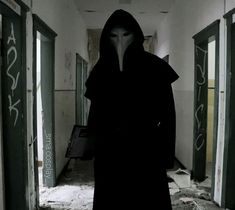 a person in a long black coat and hood standing in an empty hallway with graffiti on the walls