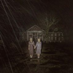 two women holding hands in front of a house at night with the light shining on them