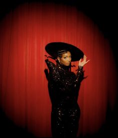 a woman in a black dress and hat posing with her hands on her head, against a red curtain