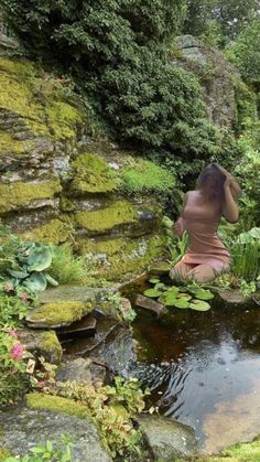 a woman sitting on top of a lush green hillside next to a small pond filled with water lilies