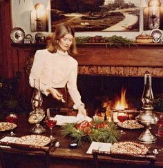 a woman standing in front of a fire place with food on the table next to her