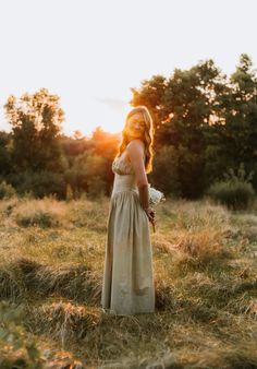 a woman in a dress standing in a field with trees behind her and the sun shining on her face