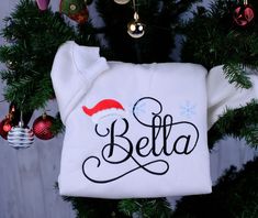 a personalized christmas sweater hanging from a tree with ornaments around it and the name bella on it