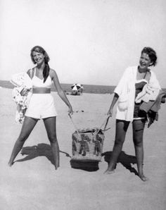 two women in bathing suits standing on the beach with their arms around each other while holding luggage