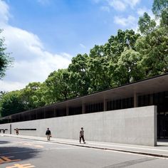 two people walking down the street in front of a concrete building with trees on both sides