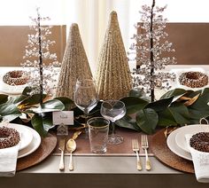 the table is set with white plates and silverware, gold napkins and trees