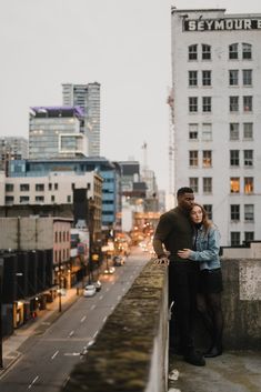 a man and woman standing next to each other on top of a bridge in front of tall buildings