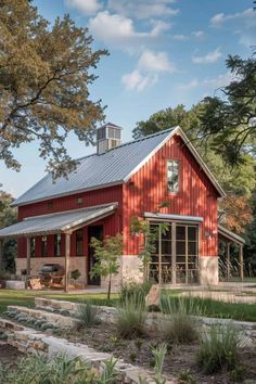 a red barn with a metal roof and two windows on the front, surrounded by trees