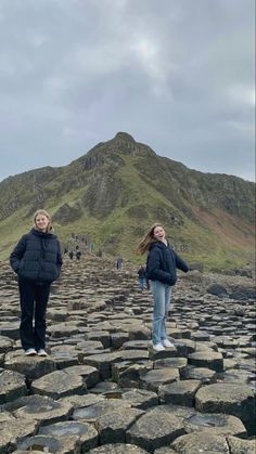 two women standing on the rocks in front of a mountain with people walking around it