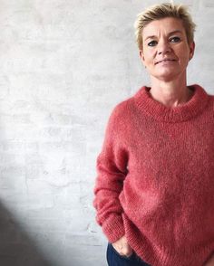 a woman standing in front of a white brick wall wearing a red sweater and jeans