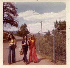 three women standing on the side of a road next to a chain link fence with trees in the background