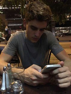 a young man sitting at a table looking at his cell phone while holding it in his hand