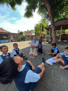 a group of children sitting on the ground in front of a woman pointing at them