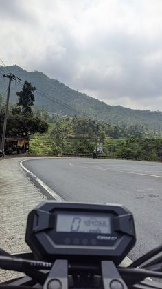 a motorcycle is driving down the road in front of some hills and trees with a clock on it's handlebars