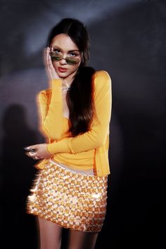 a woman in yellow shirt and gold sequin skirt holding her hand up to her face