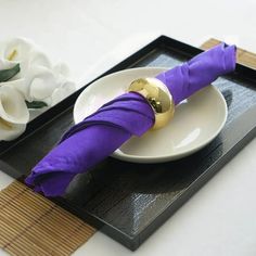 an empty plate with a purple napkin and gold ring on it next to white flowers