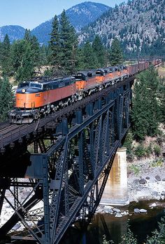 an orange train traveling over a bridge next to a forest and mountain range in the background