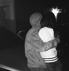 two people hugging each other while standing in front of a car on the street at night