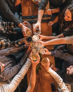 a group of people sitting around a wooden table holding glasses with drinks in them and their hands together