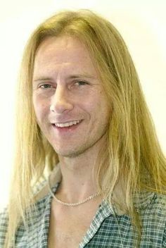 a man with long blonde hair wearing a green shirt and silver necklace smiling at the camera