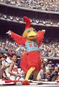 a person in a chicken costume standing on top of a baseball field while people watch from the bleachers