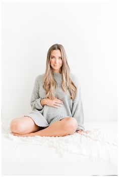 a woman sitting on top of a bed wearing a gray sweater and white pants with her legs crossed