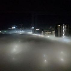 fog is covering the ground and buildings in the distance at night with lights shining on them