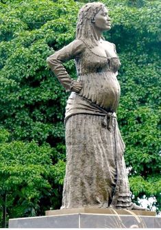 a statue of a woman standing in front of some trees and bushes with her hands on her hips