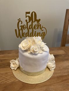 a white cake with gold lettering and flowers on top sitting on a wooden table next to a chair