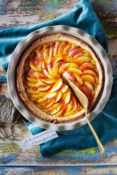 a pie with sliced peaches in it on top of a blue cloth next to a spoon