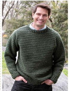 a man wearing a green sweater standing in front of a rock with his hands on his hips