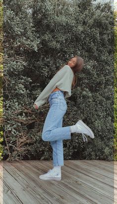 a woman standing on top of a wooden floor next to a tree and bushes with her arms in the air