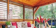 a covered porch with couches and flowers on the table in front of windows that have wooden slats over them