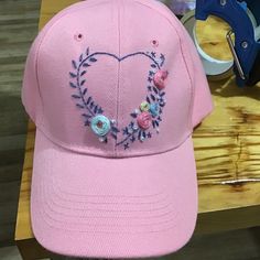 a pink baseball cap with embroidered heart and flowers on the front, sitting on a wooden table