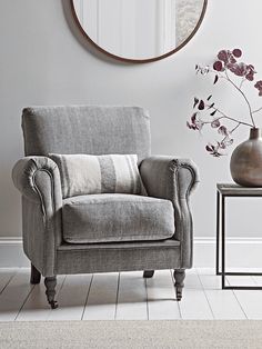 a gray chair sitting next to a vase with flowers in it and a round mirror on the wall