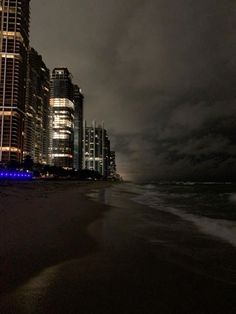 the city is lit up at night on the beach with dark clouds in the sky