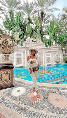 a woman standing in front of a pool wearing a white outfit and hat with palm trees behind her