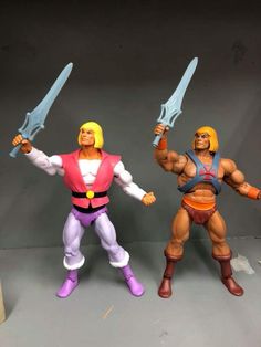 two action figures are posed next to each other on a gray surface, one is holding two swords