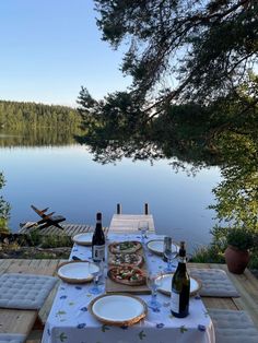 an outdoor table with plates and wine bottles on it next to a lake in the background