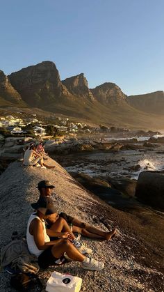 two people sitting on the beach with mountains in the backgrouund and water behind them