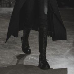 a woman walking down a runway wearing black boots and a long coat over her shoulders