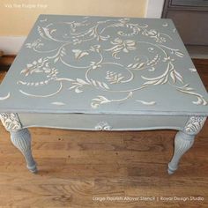 an old table painted in blue with white flowers and scrolls on the top, sitting on a hard wood floor