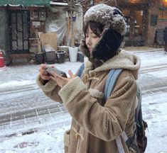 a woman standing in the snow looking at her cell phone while she is wearing a winter coat