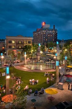 people sitting and walking around in the evening at an outdoor park with fountains, tables, and umbrellas