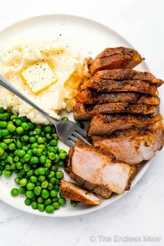 a white plate topped with meat, mashed potatoes and green peas next to a fork