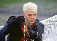 a man with blonde hair holding a microphone next to a hammock in a park