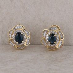 This pair of 14k yellow gold earrings have 2, 7x5 mm oval blue sapphires at the centers. There are twenty-four round brilliant diamonds that measure 0.15ctw in the first halo and sixty baguettes in the outer halo. The earrings have post and omega backs, weighing 7.4 grams together. One earring measures 14.1 mm in size. Condition: Pre-Owned *All items are thoroughly inspected for quality assurance purposes Yellow Gold Earrings, Double Halo, Sapphire Earrings, Yellow Gold Earring, Fine Jewellery Earrings, Quality Assurance, Jewelry Earrings Studs, Round Brilliant, Blue Sapphire