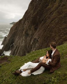 a man and woman sitting on the grass by the ocean with cliffs in the background