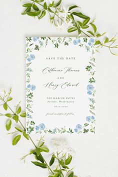 a wedding card with blue flowers and greenery on the front, next to some green leaves