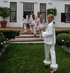 a man dressed in white standing on the grass with other people behind him and two girls walking by